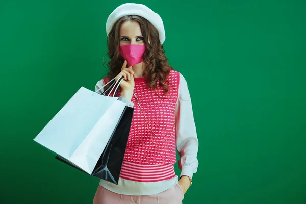 Covid 19パンデミックの間の生活 ピンクの医療マスクと緑の背景に隔離されたショッピングバッグを持つ白いベレー帽の高価なエレガントな女性買い物客 — ストック写真