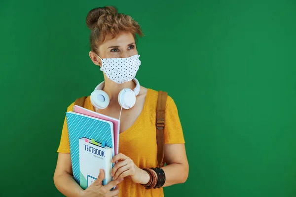 在大肠病毒大流行期间的生命 身穿黄色衬衫的女学生 带着课本 笔记本 医疗面罩 白色耳机和背包 望着绿色背景隔离开的复印空间 — 图库照片