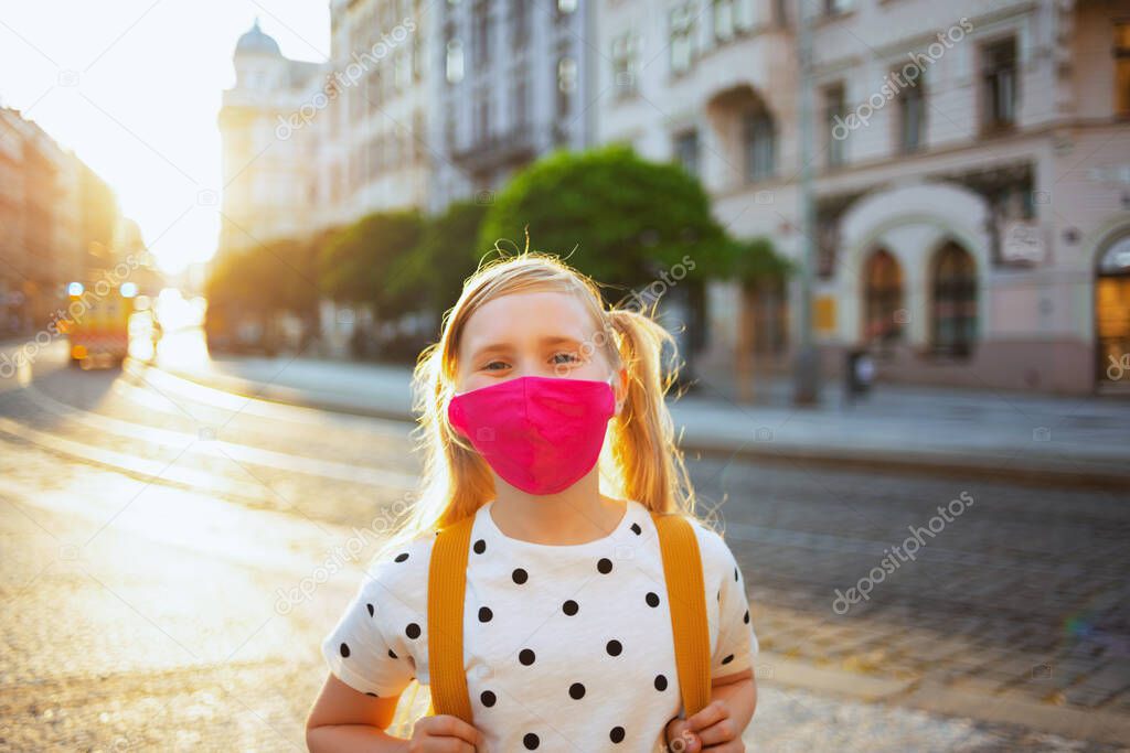 Life during coronavirus pandemic. Portrait of modern child in white polka dot blouse with pink mask and yellow backpack coming back from school outside.