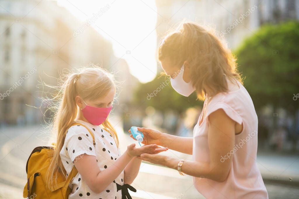 Life during covid-19 pandemic. young mother and daughter with masks and yellow backpack disinfecting hands with sanitiser and getting ready for school outside.