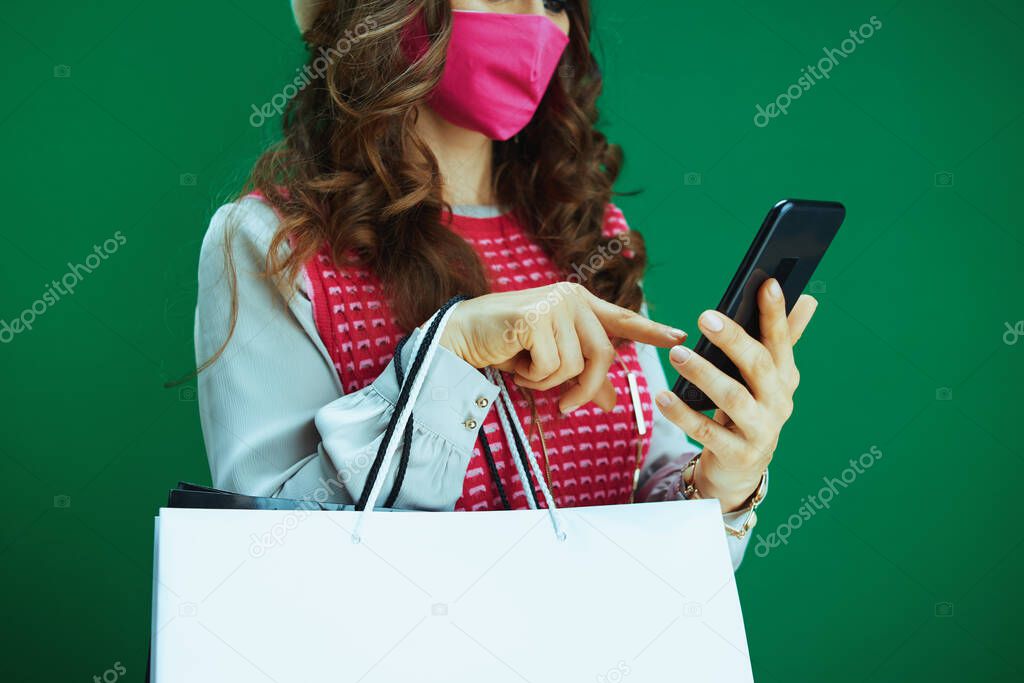 Life during coronavirus pandemic. Closeup on woman shopper in white beret with pink medical mask and shopping bags buying online on a smartphone on green background.