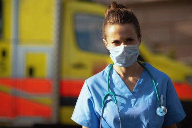 Covid-19 salgını. Dışarıda ambulansın yanında steteskoplu ve tıbbi maskeli modern tıp doktoru portresi..