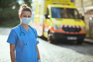 Covid-19 salgını. Dışarıda ambulansın yanında steteskop ve tıbbi maske takan modern sağlık görevlisi kadın portresi..
