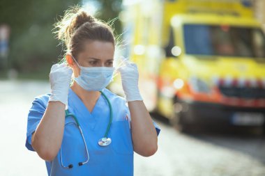 Covid-19 salgını. Dışarıda ambulansın yanında steteskop ve tıbbi maske takan üniformalı modern tıp doktoru kadın..