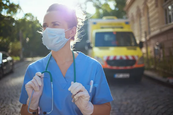 验尸官大流行病 身穿制服 戴着听诊器和医疗面罩的现代辅助医疗妇女 向外面靠近救护车的地方望去 — 图库照片
