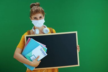 Covid-19 salgını sırasında hayat. Ders kitapları, not defterleri, tıbbi maske, beyaz kulaklıklar ve yeşil arka planda izole edilmiş boş tahta gösteren sırt çantalı genç bayan öğrenci..