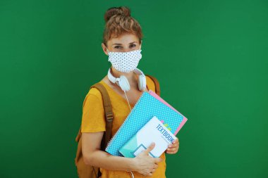 Koronavirüs salgını sırasında hayat. Sarı tişörtlü gülümseyen genç öğrencinin portresi ders kitapları, defterler, tıbbi maske, beyaz kulaklık ve yeşil arka planda sırt çantası..