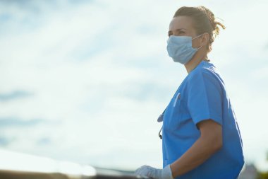 Covid-19 salgını. Kendine güvenen, doktor önlüklü, tıbbi maskeli ve lastik eldivenli bir kadın şehrin dışında gökyüzüne karşı mesafeye bakıyor..