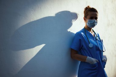 Koronavirüs salgını. Şehir dışında duvarın yanında steteskop, tıbbi maske ve lastik eldiven takan yorgun, modern tıbbi pratisyen kadın..