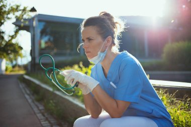 Covid-19 salgını. Steteskoplu ve tıbbi maskeli modern kadın kliniğin yanında oturuyor..