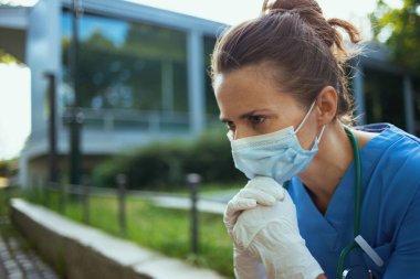 Koronavirüs salgını. Steteskop ve tıbbi maskeli, önlüklü, mutsuz, modern tıp doktoru kadın kliniğin dışında oturuyor..