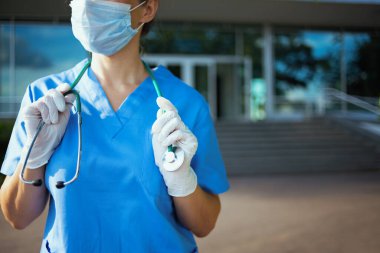 Koronavirüs salgını. Steteskop ve tıbbi maskeli, önlüklü, kendine güvenen tıbbi pratisyen kadın kliniğin yakınında..