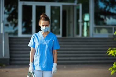 Koronavirüs salgını. Steteskop ve tıbbi maskeli, önlüklü, mutsuz, modern tıp uzmanı kadın kliniğin yakınında..