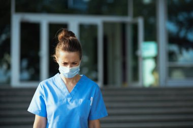 Covid-19 salgını. Hüzünlü modern tıp pratisyeni kadın kliniğin önünde tıbbi maske takıyor..