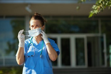 Koronavirüs salgını. Steteskop ve tıbbi maske takmış modern tıp uzmanı kadın hastanenin yakınındaki bir yere bakıyor..