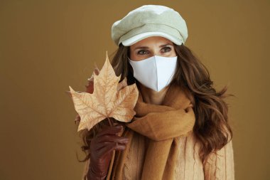 Koronavirüs salgını sırasında hayat. Beyaz tıbbi maskeli, sarı sonbahar akçaağaç yapraklı, eşarplı düşünceli genç ev hanımı bej arka plandaki mesafeye bakıyor..
