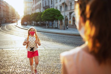 Koronavirüs salgını sırasında hayat. Maskeli ve sarı sırt çantalı zarif anne ve okul çocuğu okuldan dönüyorlar..