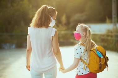 Covid-19 salgını sırasında hayat. Arkadan, maskeli, sarı sırt çantalı ve okul dezenfektanlı genç bir anne ve çocuğun okuldan çıkarken görülmüş..