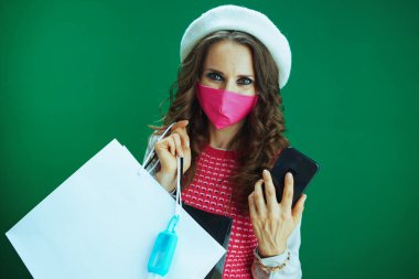 Beyaz bereli, pembe maskeli, alışveriş çantalı ve yeşil arka planda izole edilmiş akıllı telefondan internetten alan bir antibakteriyel ajanın son moda öğrenci portresi..