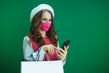 Covid-19 salgını sırasında hayat. Pembe kolsuz gömlekli şık bir kadının portresi. Pembe tıbbi maskeli ve alışveriş çantalı. Çevrimiçi bir dükkanda yeşil arka planda akıllı telefondan bakıyorlar..