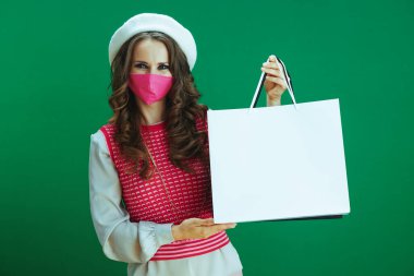 Koronavirüs salgını sırasında hayat. Pembe kolsuz gömlekli mutlu genç kadın portresi. Pembe sağlık maskesi ve yeşil arka planda izole edilmiş alışveriş çantaları..