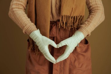Hello autumn. Closeup on middle aged woman in scarf with leather gloves showing heart shaped hands isolated on beige background. clipart