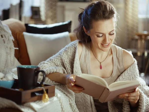 幸せな若いです女性で編んだ居心地の良いカーディガンとともにトレイとカップ読書本で現代の家で晴れた秋の日 — ストック写真