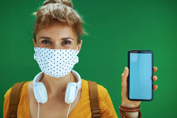 Life during covid-19 pandemic. Portrait of young student woman in yellow shirt with medical mask, white headphones and backpack showing smartphone blank screen on green background.