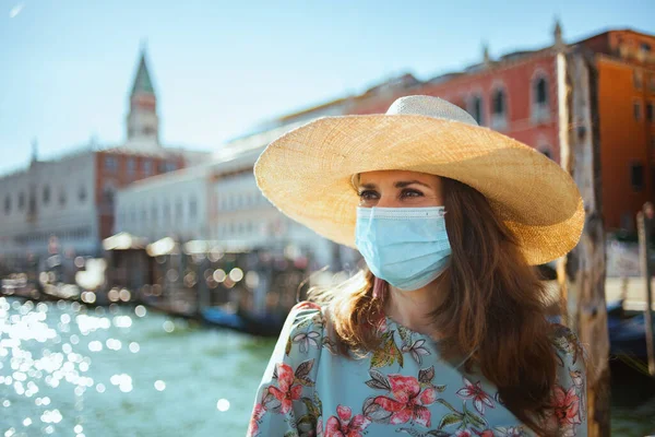Travel during covid-19 pandemic. modern middle aged traveller woman in floral dress with medical mask having walking tour near San Marco square on embankment in Venice, Italy.