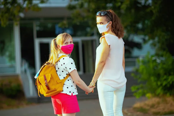 Koronavirüs salgını sırasında hayat. Modern anne ve okul çocuğu maskeli ve sarı sırt çantalı açık havada okula gidiyor..