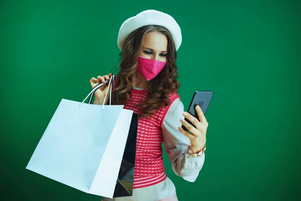 Covid 19パンデミックの間の生活 ピンクの医療マスクとショッピングバッグを持つピンクのノースリーブシャツの若い学生緑の背景にスマートフォンでオンラインショップを閲覧 — ストック写真