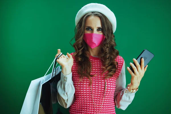 ピンクの医療用マスクとショッピングバッグを持つ白いベレー帽の流行の女性の買い物客の肖像は 緑の背景にスマートフォン上の電子商取引サイト上でオンラインショッピングを作る — ストック写真