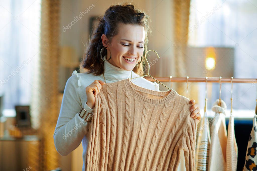 happy elegant woman in white sweater and skirt in the modern house in sunny winter day choosing sweaters hanging on copper clothes rack.