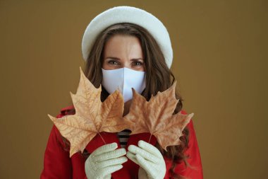 Koronavirüs salgını sırasında hayat. Kahverengi arka planda sarı sonbahar akçaağaç yaprağı olan kırmızı ceketli genç bir kadının portresi..