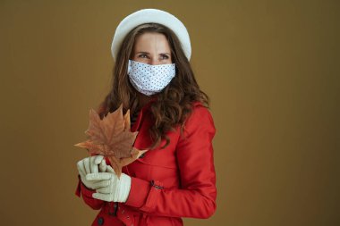 Covid-19 salgını sırasında hayat. Sarı sonbahar akçaağaç yaprağıyla kırmızı ceketli hayalperest modern ev kadını kahverengi arka planda izole edilmiş mesafeye bakıyor..