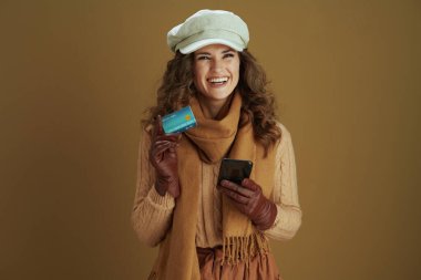 Merhaba Kasım ayı. Akıllı telefon, deri eldiven ve kredi kartıyla elektronik ticaret sitesinde kahverengi arka planda alışveriş yapan mutlu stil sahibi bir kadın..