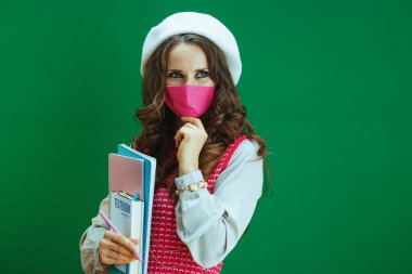 Covid-19 salgını sırasında hayat. Pembe kolsuz gömlekli, ders kitabı, not defteri ve yeşil üzerine izole edilmiş pembe tıbbi maskeli düşünceli genç kadın..