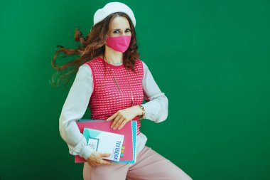 Covid-19 salgını sırasında hayat. Pembe kolsuz gömlekli neşeli modern kadın ders kitapları, defterler ve pembe tıbbi maske ile yeşil arka planda izole edilmiş..