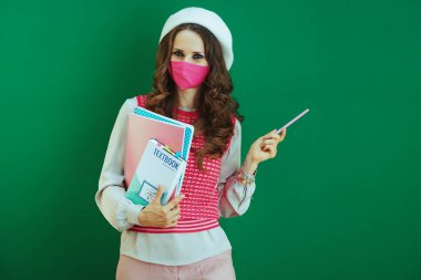 Covid-19 salgını sırasında hayat. Ders kitapları, not defterleri ve pembe tıbbi maskeli beyaz bereli şık bir kadın yeşil arka plandaki bir şeyi işaret ediyor..