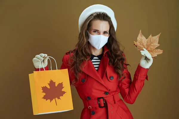 Life during coronavirus pandemic. happy trendy woman in red coat with yellow autumn maple leaf and paper shopping bag on beige background.