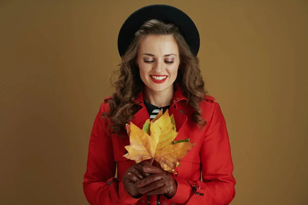 11月だ 幸せな現代中高年女性で赤コートと黒ベレー帽で革手袋と黄色の秋のカエデの葉はベージュの背景に隔離 — ストック写真