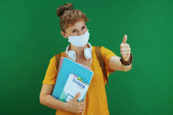 Covid 19大流行病期间的生活 穿着黄色衬衫的现代女学生 带着课本 笔记本 医疗面罩 白色耳机和背包 大拇指贴在绿色背景上 — 图库照片