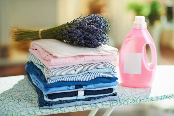 pile of washed clothes, pink bottle of fabric softener and bunch of lavender on ironing board.