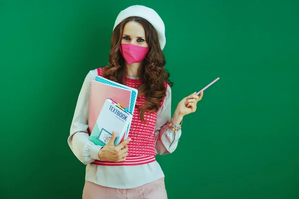 Covid 19大流行病期间的生活 穿着白色贝雷帽的时髦女人 带着课本 笔记本和粉色医疗面罩 指着绿色背景的东西 — 图库照片
