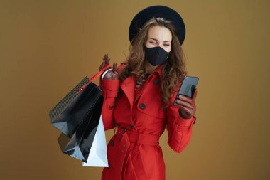 Merhaba sonbahar. Siyah maskeli ve kağıt çantalı moda bir kadın bronz arka planda akıllı telefon kullanarak kısa mesaj gönderiyor..