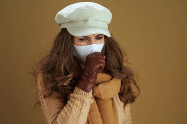 Koronavirüs salgını sırasında hayat. Beyaz tıbbi maskeli, bronz arka planda öksüren 40 yaşında zarif bir kadın..