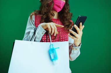 Pembe kolsuz gömlekli, pembe tıbbi maskeli, alışveriş çantalı ve antiseptik alışverişi yapan öğrencilere yakın çekim yeşil arka plandaki bir akıllı telefondan e-ticaret sitesinde.