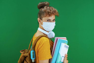 Koronavirüs salgını sırasında hayat. Yeşil arka planda izole edilmiş sarı tişörtlü, ders kitapları, defterler, tıbbi maske, beyaz kulaklık ve sırt çantalı modern kadın öğrenci..