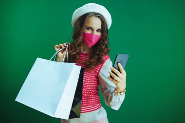 Covid-19 salgını sırasında hayat. Pembe kolsuz gömlekli şık bayan alışverişçi. Pembe sağlık maskesi ve alışveriş torbaları yeşil arka planda akıllı telefondan satın alıyor..