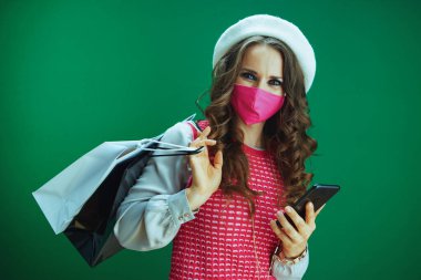 Covid-19 salgını sırasında hayat. Beyaz bereli mutlu genç bir kadının portresi. Pembe tıbbi maskeli ve alışveriş çantalı. Çevrimiçi bir telefondan yeşil arka planda geziniyor..
